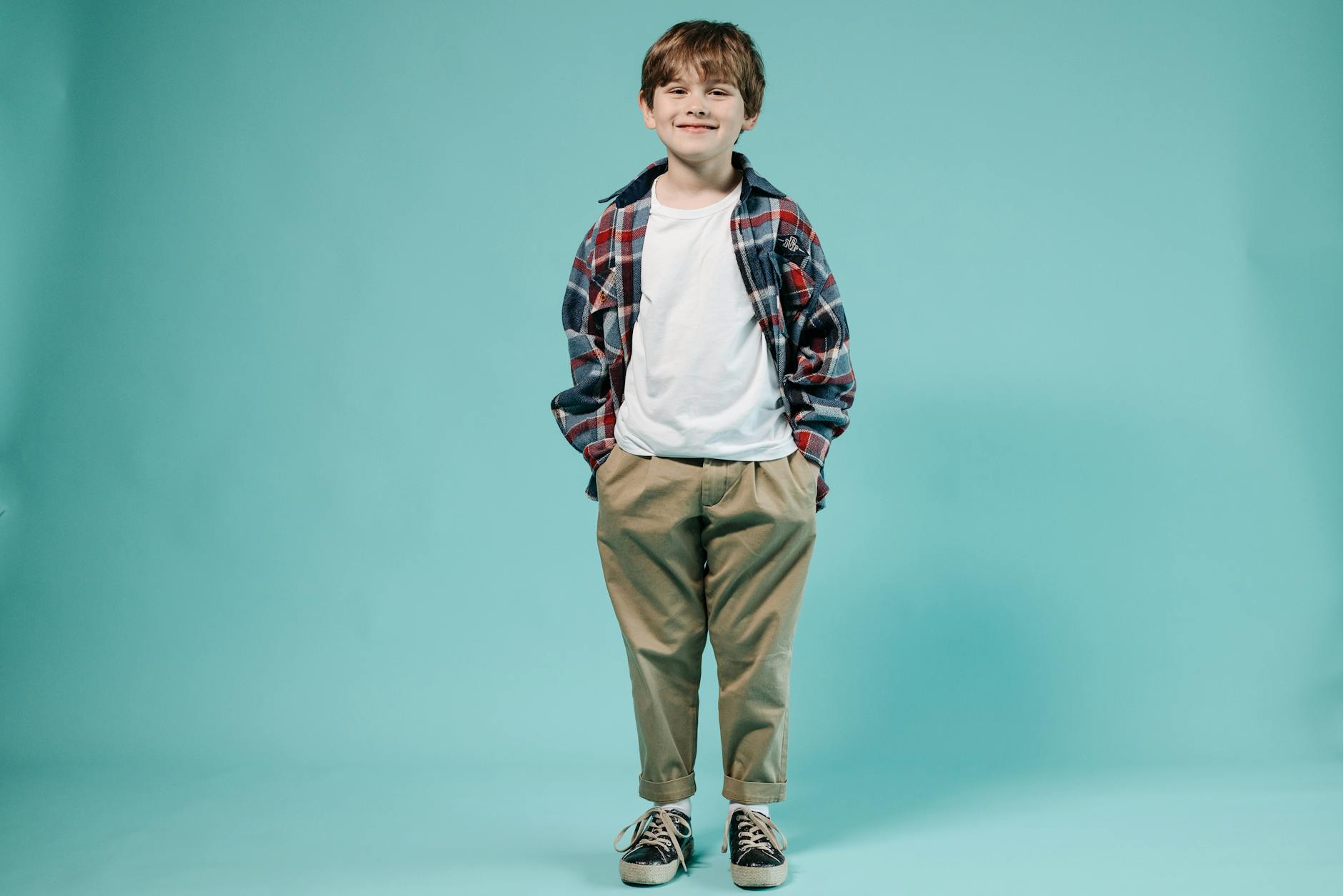 Uśmiechnięty chłopiec w koszuli w kratę z rękami w kieszeniach na niebieskim tle