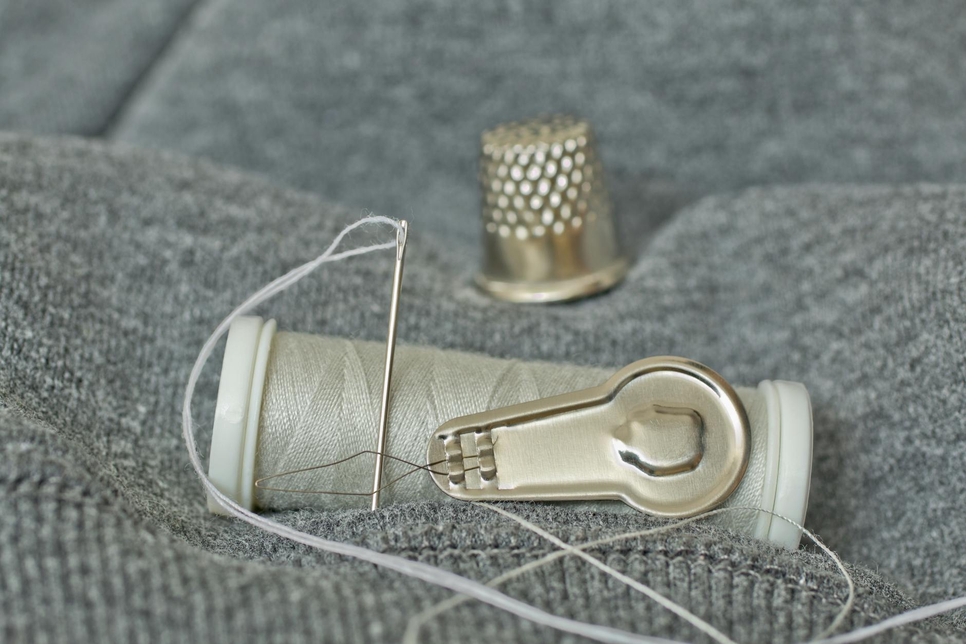 Zbliżenie na igłę, szpulkę nici i naparstek na szarym materiale